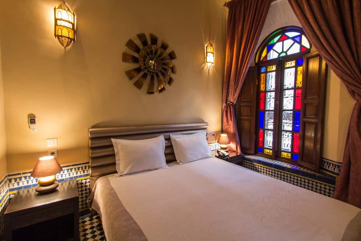 A hotel room with a bed, two bedside lamps, a wall clock, a stained glass window, and patterned tile flooring.