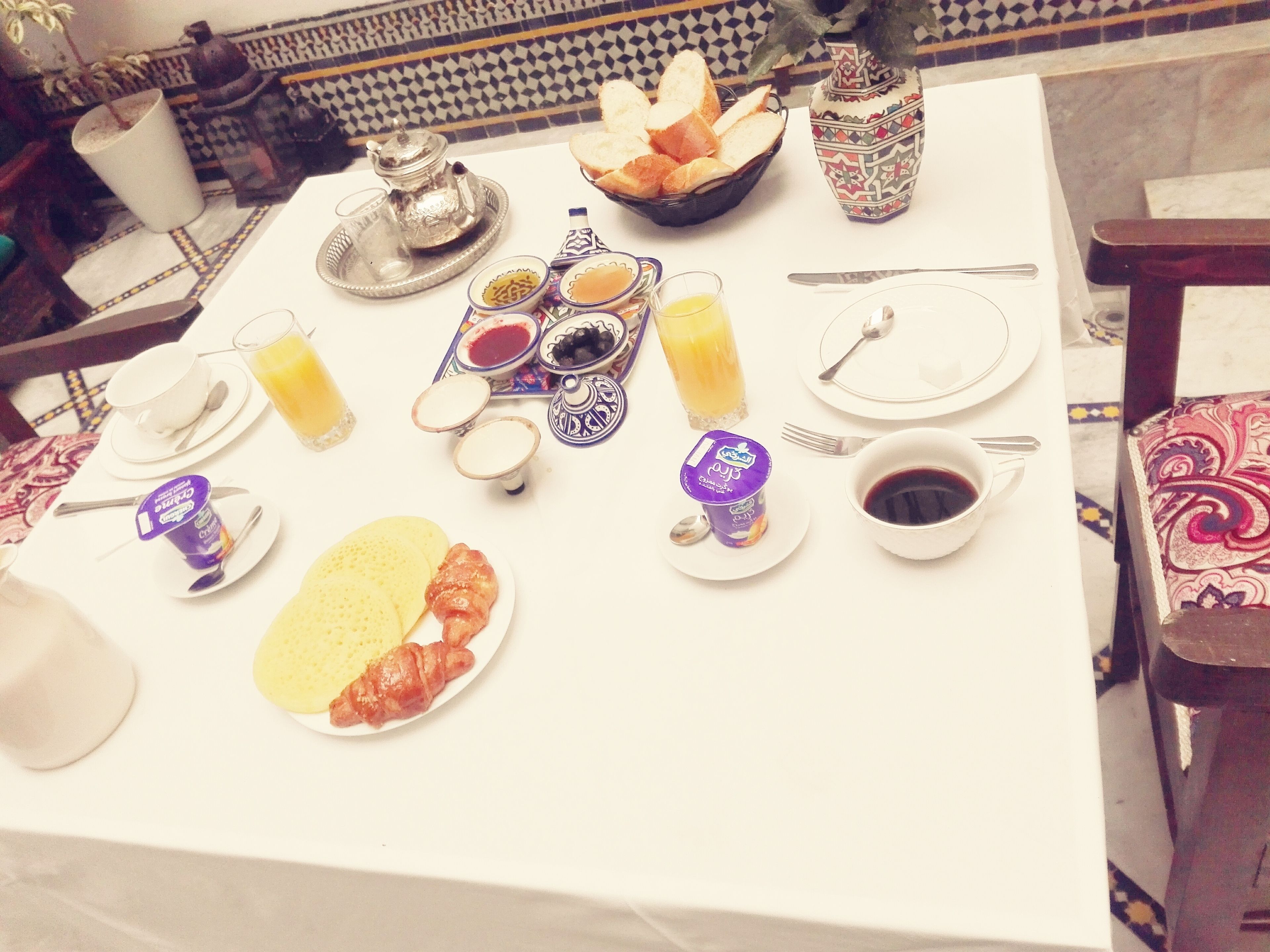 A breakfast table set with plates of food, including croissants and a bowl of bread, along with various condiments and beverages.