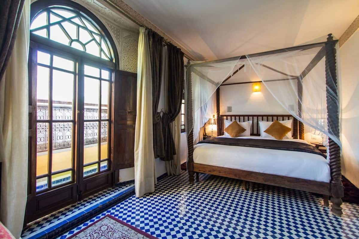 A room with a checkered floor, a bed with a canopy, and a view through a window with a decorative frame.