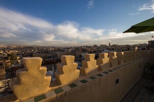 Terraço no telhado com vista para a cidade, apresentando uma série de estruturas de parede bege e geométricas, e um guarda-sol verde.