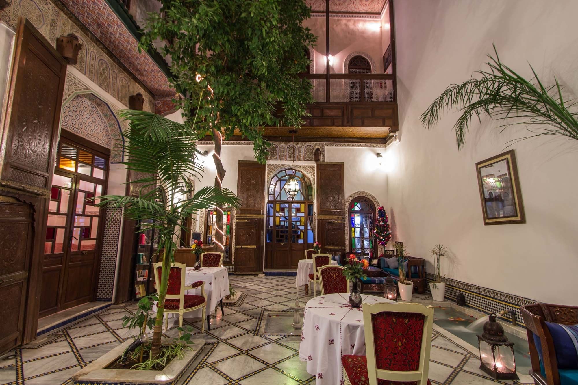 Un patio interior con techo alto, puertas de madera ornamentadas y un área central con mesas y sillas.