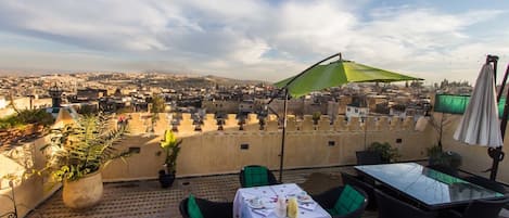 A terrace with a table set for two, a green umbrella, and a view of a cityscape.