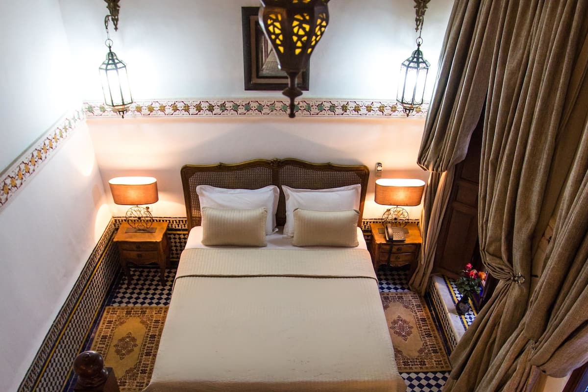 A bedroom with a bed, two bedside tables, a hanging light fixture, a patterned rug, and a checkered floor.