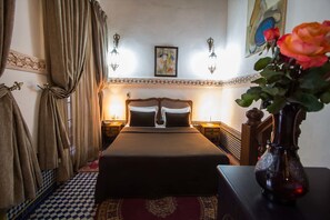 Une chambre d’hôtel avec un sol à carreaux, un lit avec une tête de lit, une table de chevet, un vase avec des fleurs et un tableau au mur.