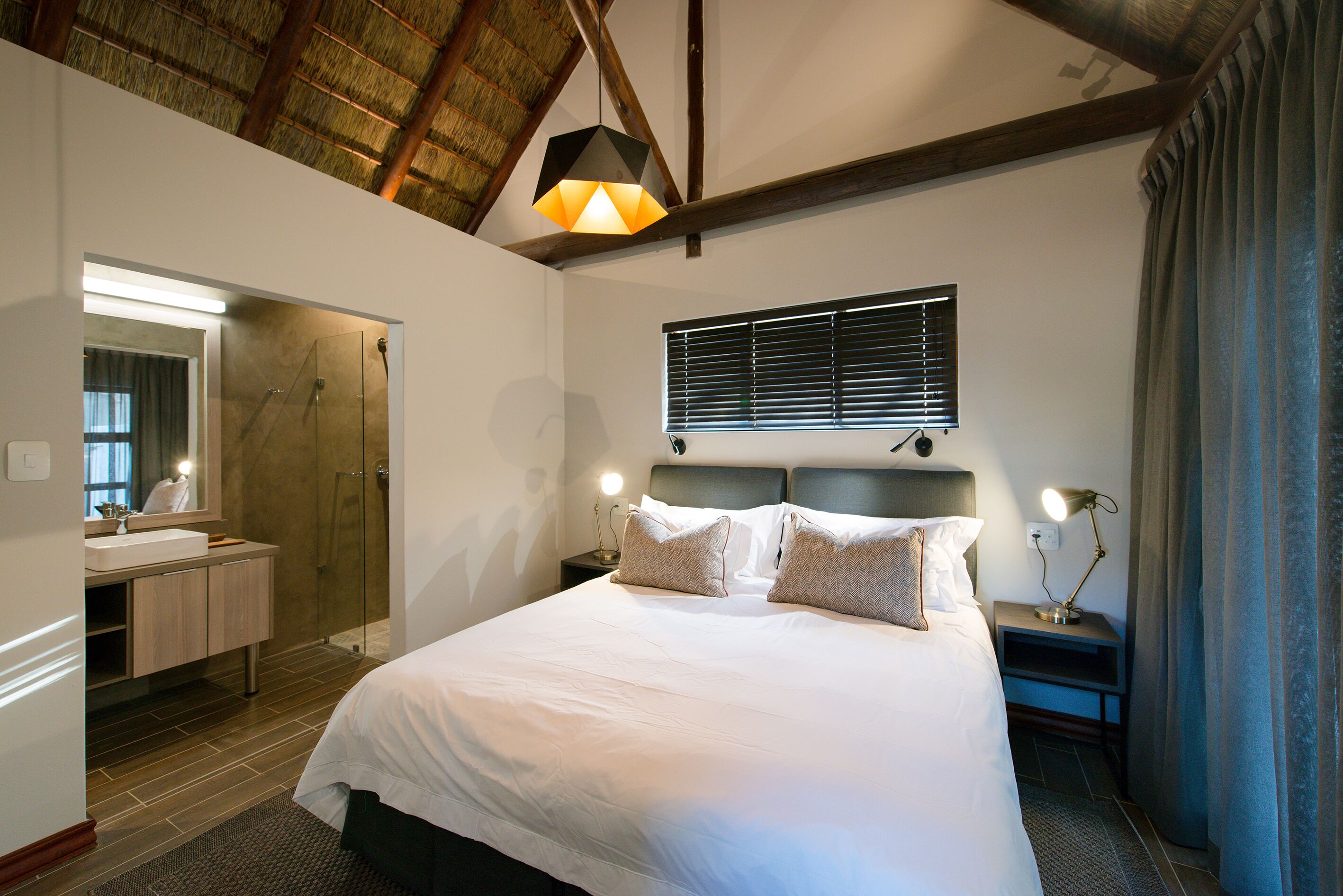 A bedroom with a large bed, a nightstand with a lamp, a bedside table, a mirror, a shower, and a window with blinds.
