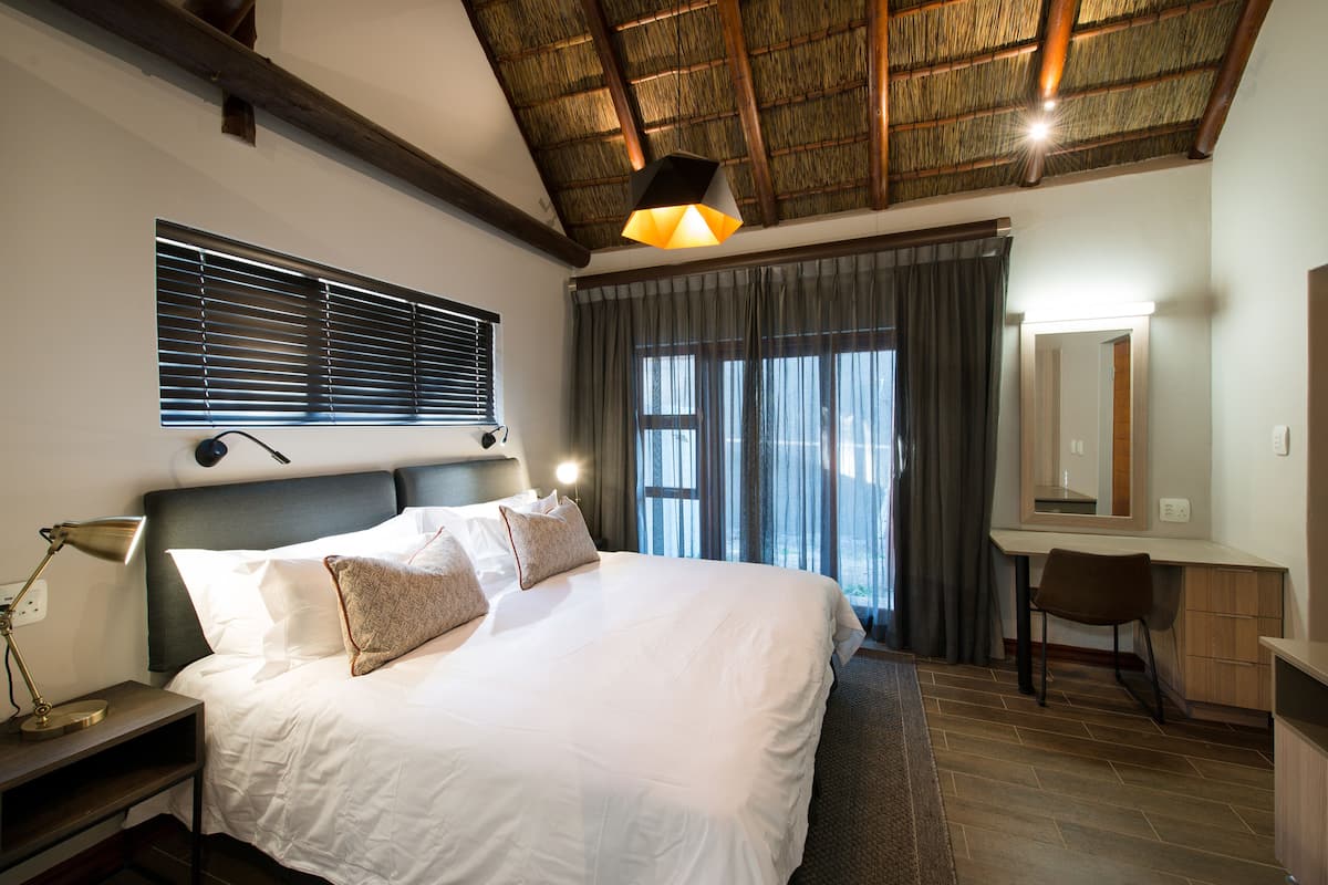 Une chambre d’hôtel avec un grand lit, un bureau, une chaise, une lampe de bureau, un miroir et une fenêtre avec des stores.