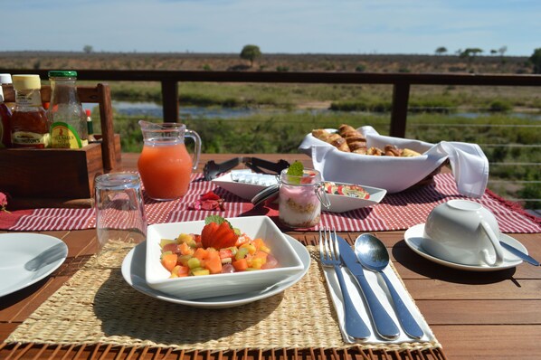 A breakfast setup with a fruit bowl, croissants, and a beverage on a wooden table with a scenic view.