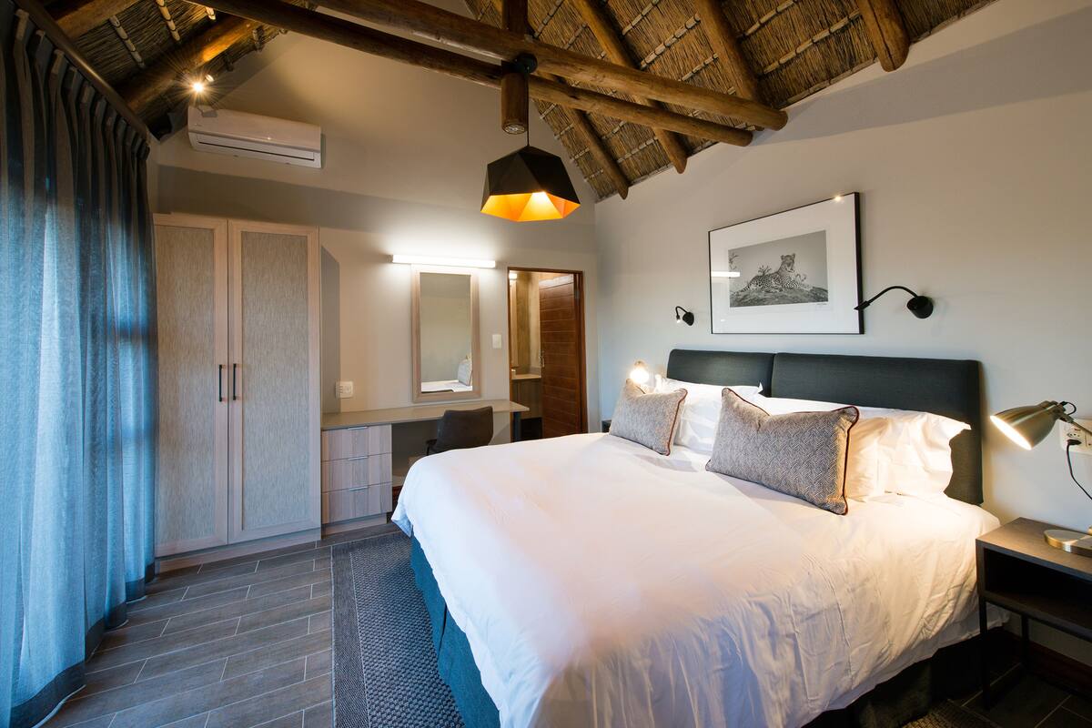 Une chambre d’hôtel avec un grand lit, un bureau, une chaise, une armoire et un plafond avec des poutres apparentes.