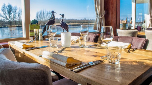 A dining table set with wine glasses, cutlery, and a menu, overlooking a lake with birds.