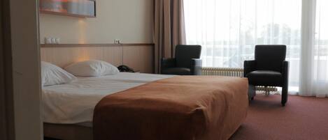 A hotel room with a bed, two chairs, a radiator, and a window with curtains.