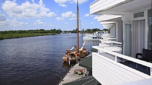 A riverside building with a balcony overlooking a river, a sailboat docked nearby, and a row of boats in the background.
