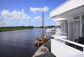 Um edifício às margens do rio com varanda que dá para a água, um veleiro atracado perto e uma fileira de barcos ao fundo.