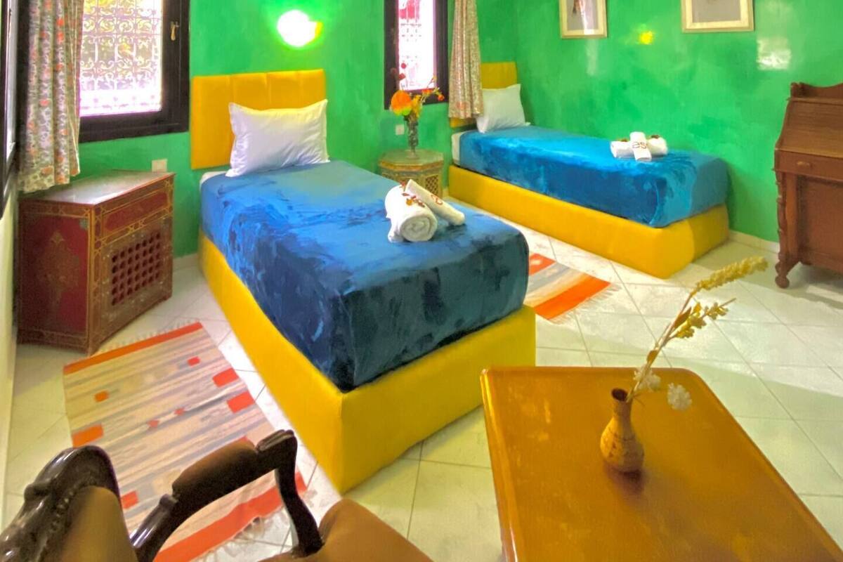 Habitación de hotel con dos camas, una mesita, una silla y una ventana con cortinas.