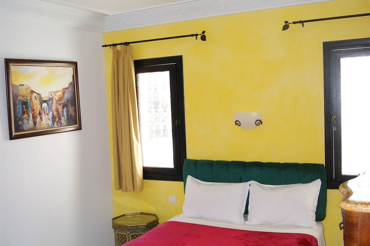 Una habitación con una cama, un cuadro, una ventana con cortinas y una lámpara en el techo.