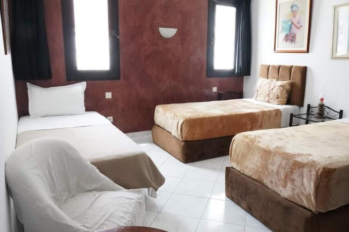 Habitación de hotel con dos camas, una silla y una mesita pequeña. Cuenta con dos ventanas con cortinas, un reloj de pared y cuadros en la pared.