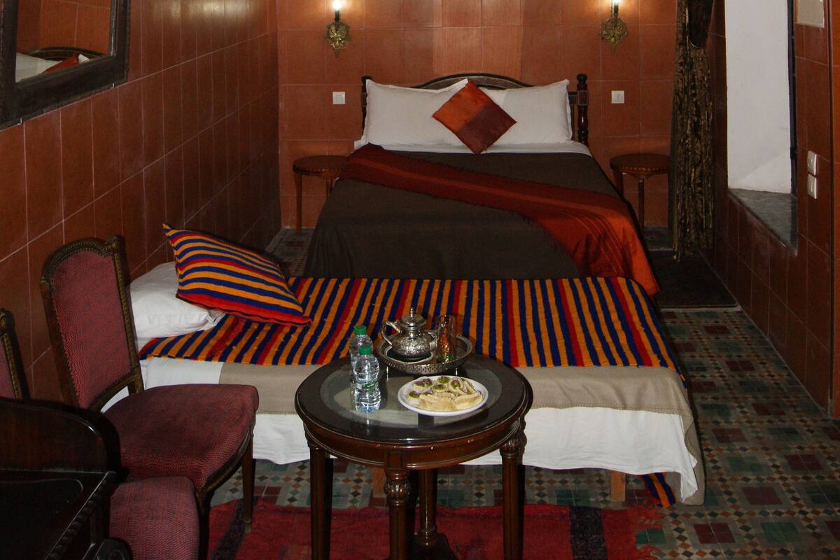 Una habitación de hotel con cama, una silla, una mesa con una botella y un plato, una araña y un espejo.
