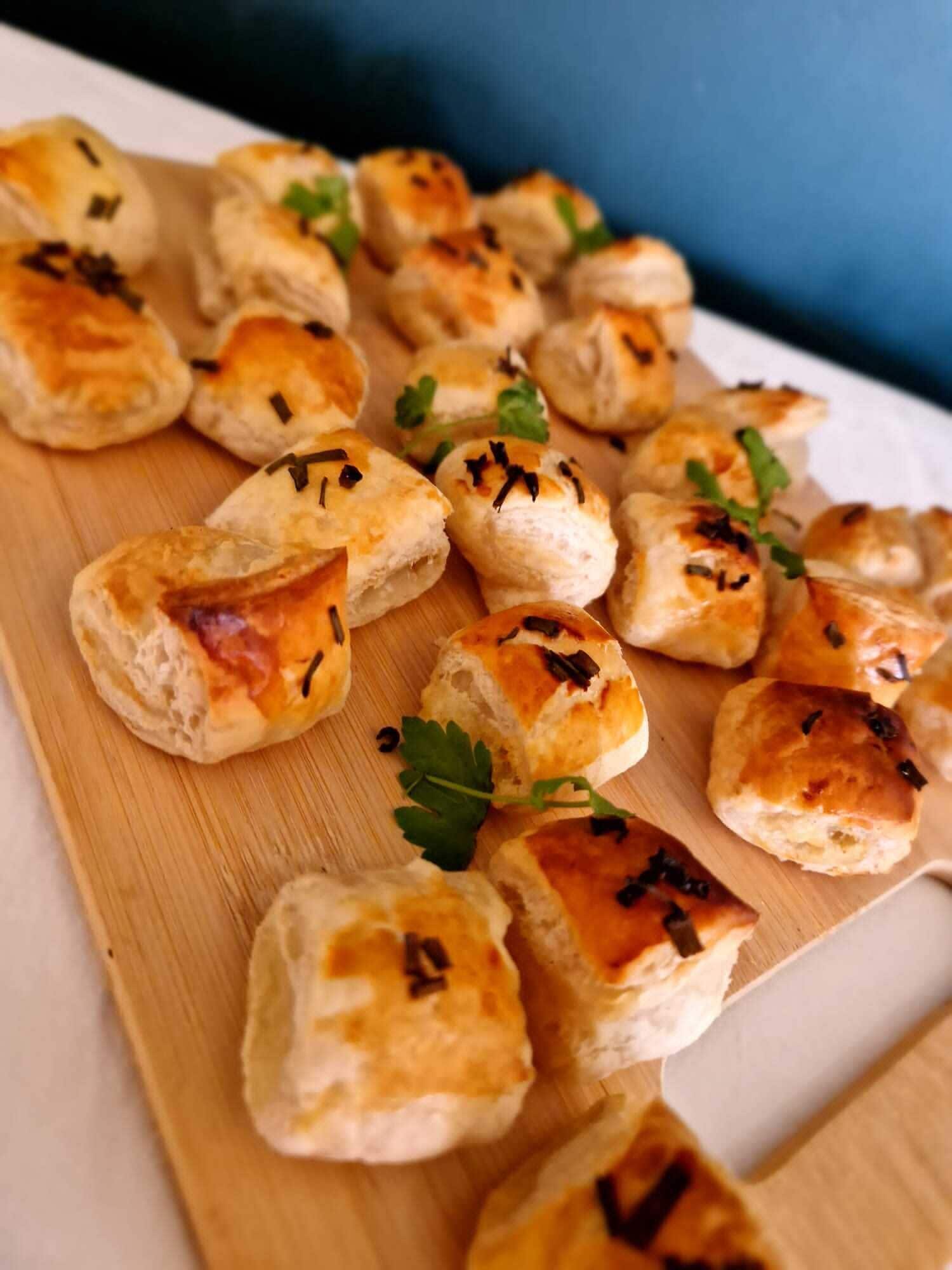 A wooden board with several small, golden-brown pastries garnished with herbs and black specks, likely spices, on a white surface.