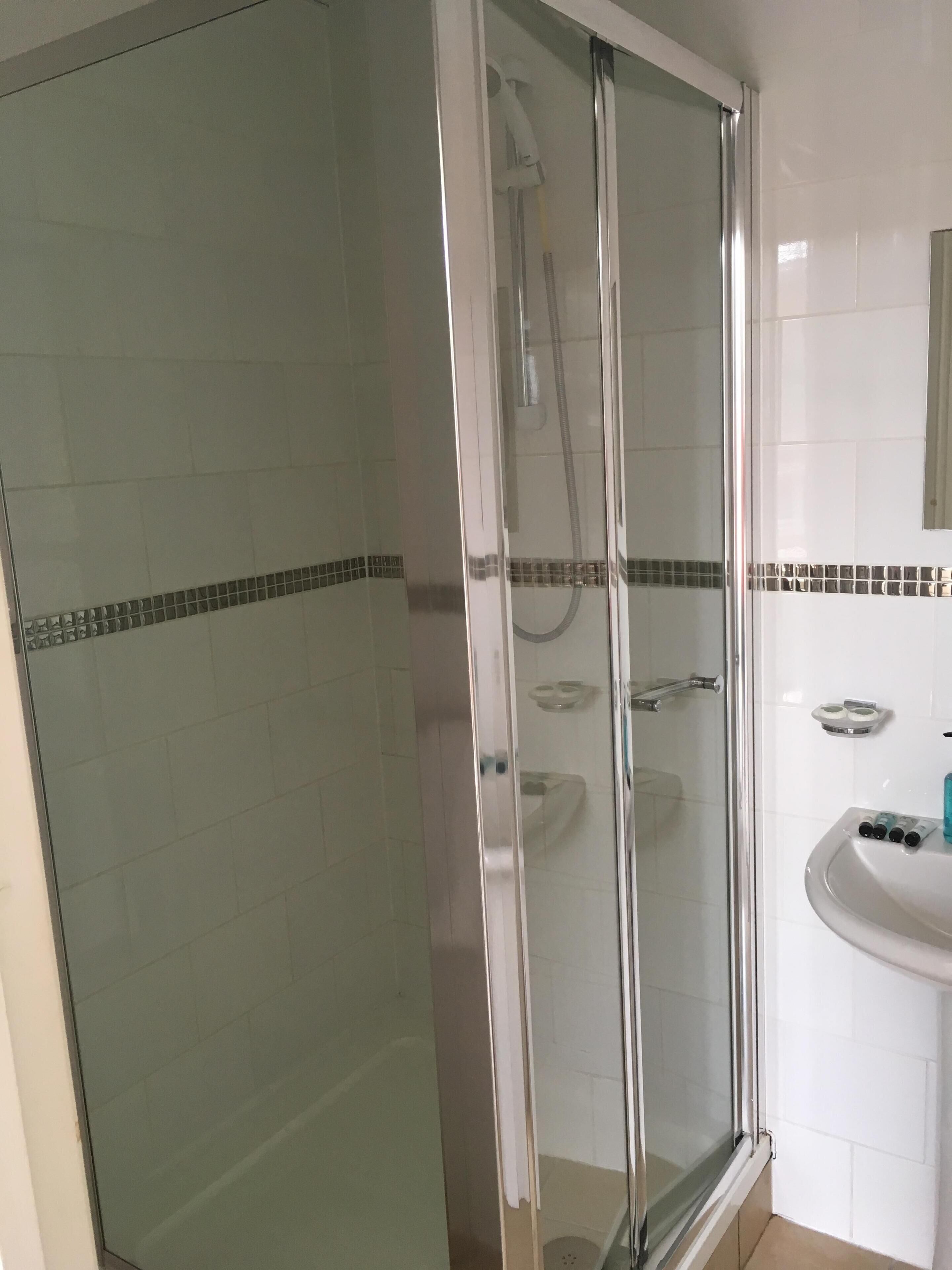 Eine Duschkabine mit Glas, eine weiße Fliesenwand und ein kleines Waschbecken mit Pflegeprodukten.