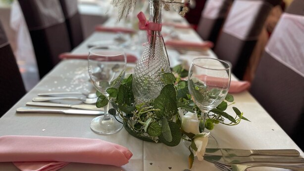 Una mesa de comedor adornada con una botella de vidrio transparente, rosas blancas y follaje verde.