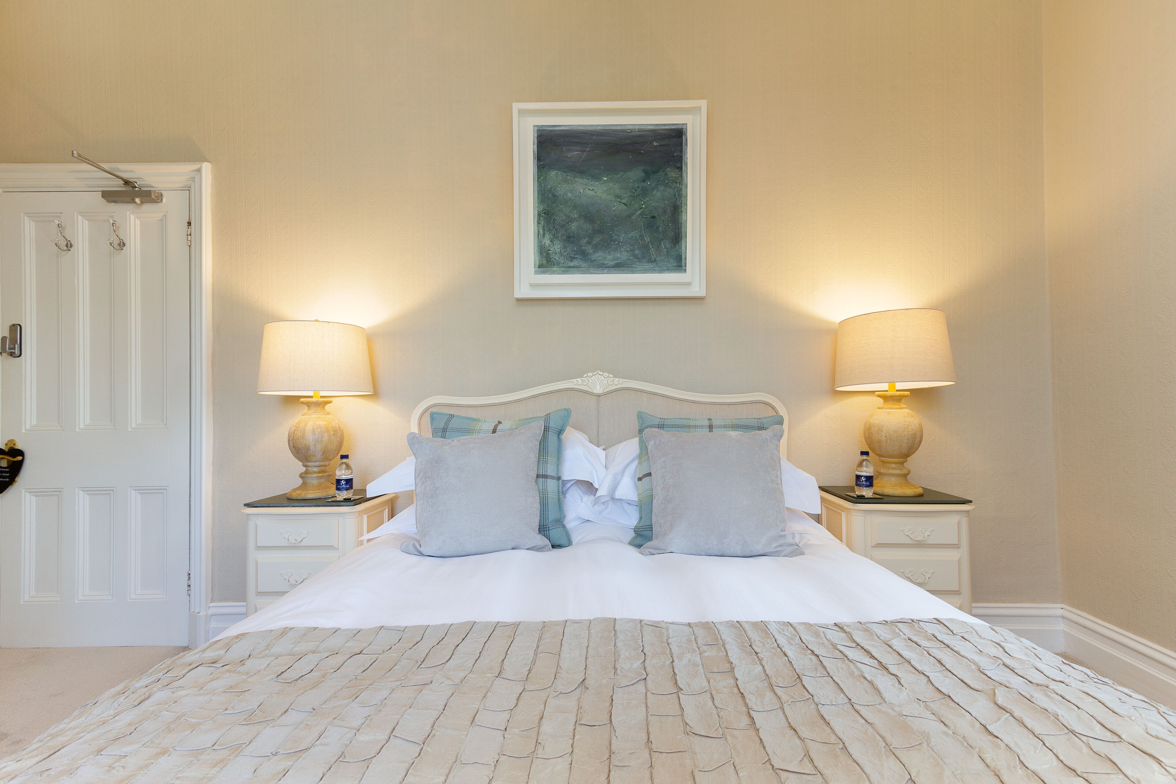 Un lit bien fait, avec une tête de lit blanche, deux tables de chevet avec des lampes, une œuvre d’art encadrée au-dessus du lit et une porte blanche sur le côté gauche.