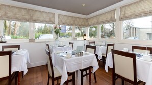 A dining area with tables set for a meal, large windows with curtains, and a view of the outdoors. - Glencree (Windermere)