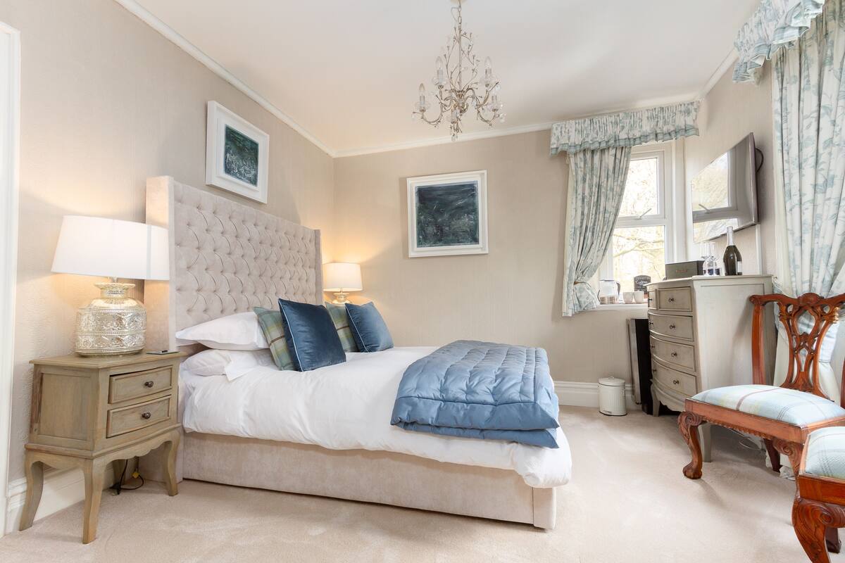 A bedroom with a large bed, bedside tables, a dresser, a chair, a chandelier, and artwork on the walls.