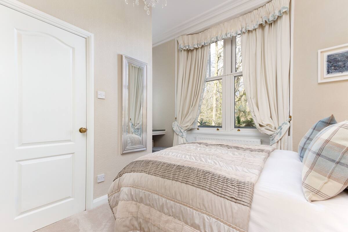 A bedroom with a bed, a window with curtains, a mirror, and a door.