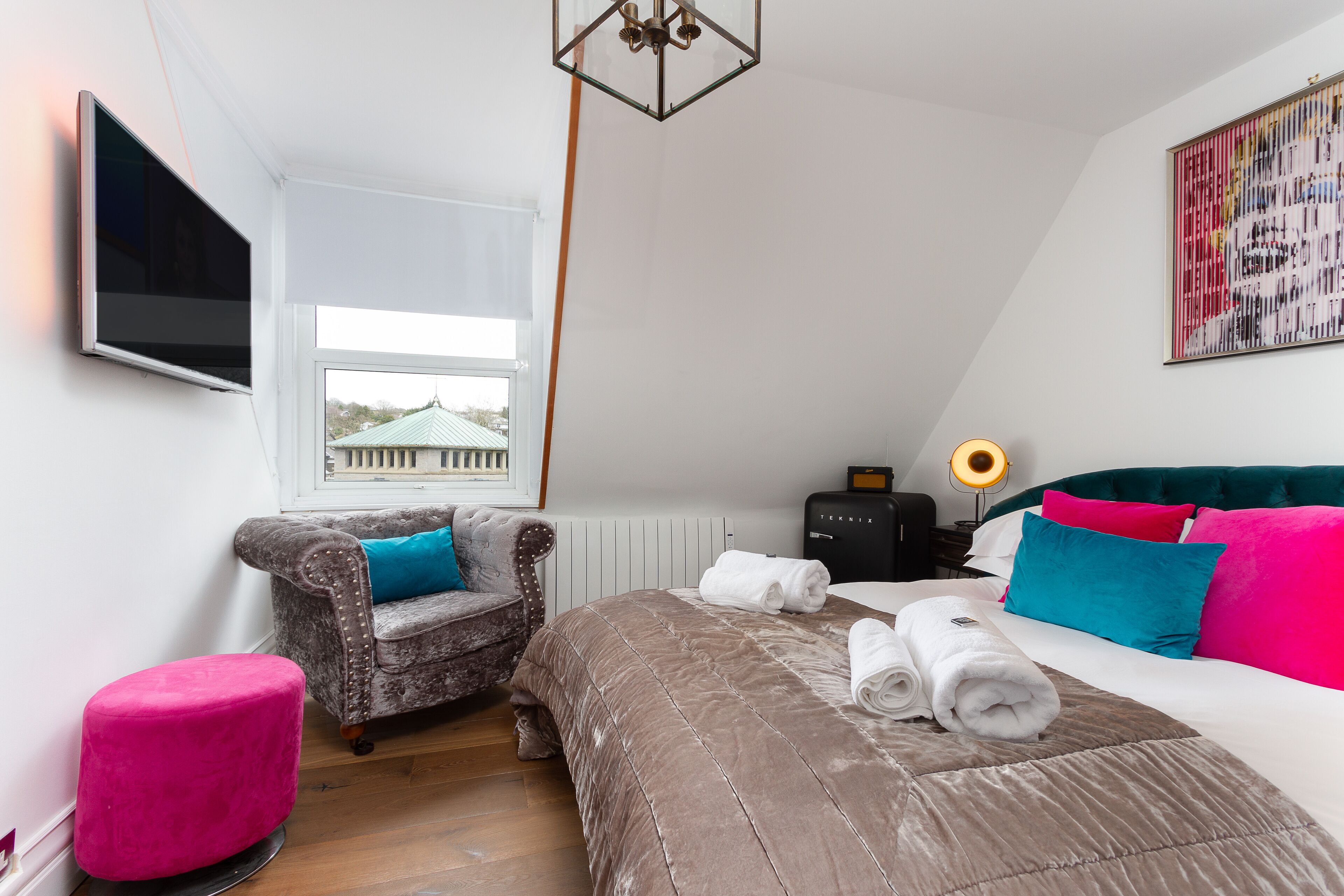 A bedroom with a bed, a chair, a pink ottoman, a television, and a window with a view of a building.