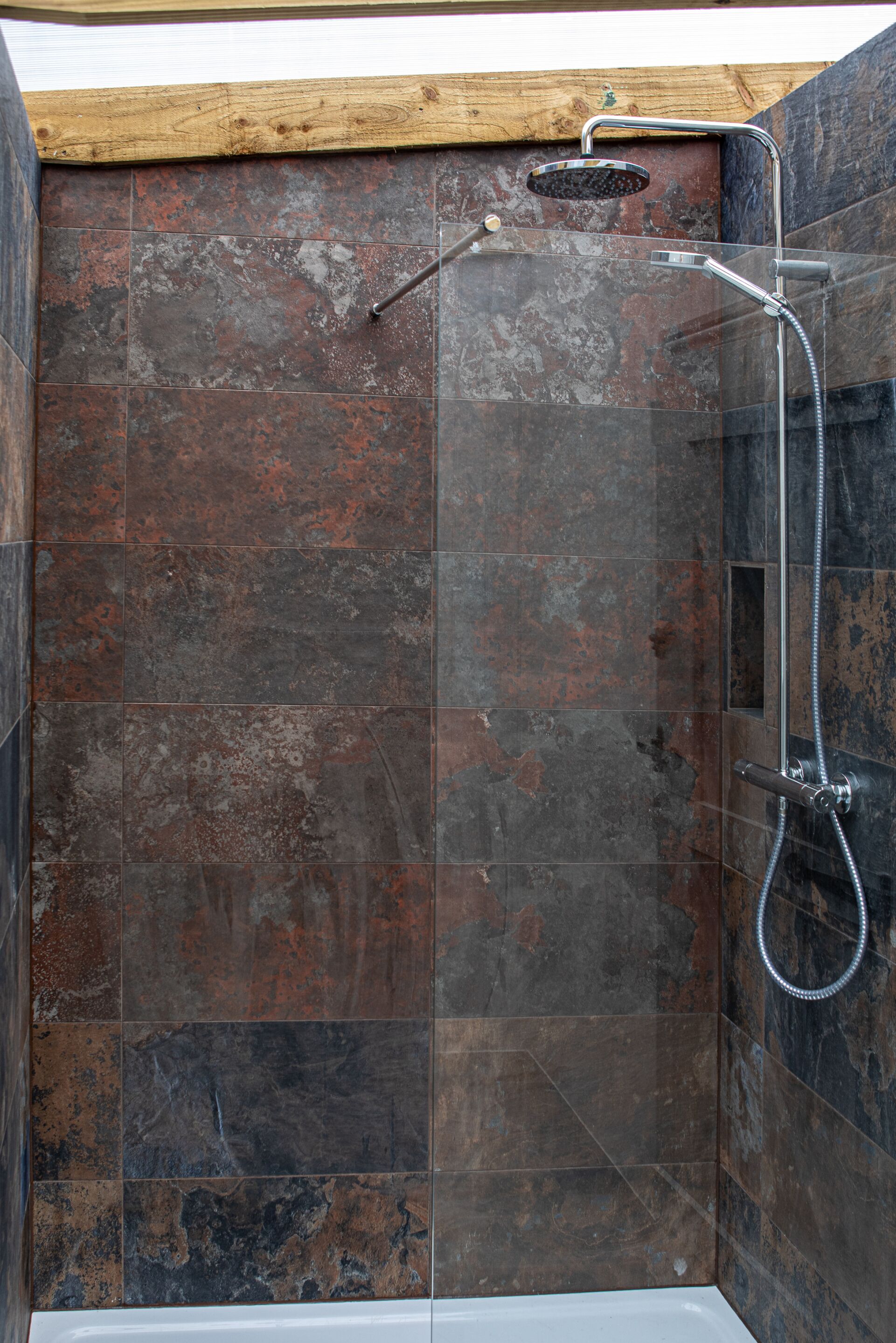 A shower with a handheld showerhead and a wall-mounted faucet, set in a room with dark, textured tiles and a wooden ceiling.