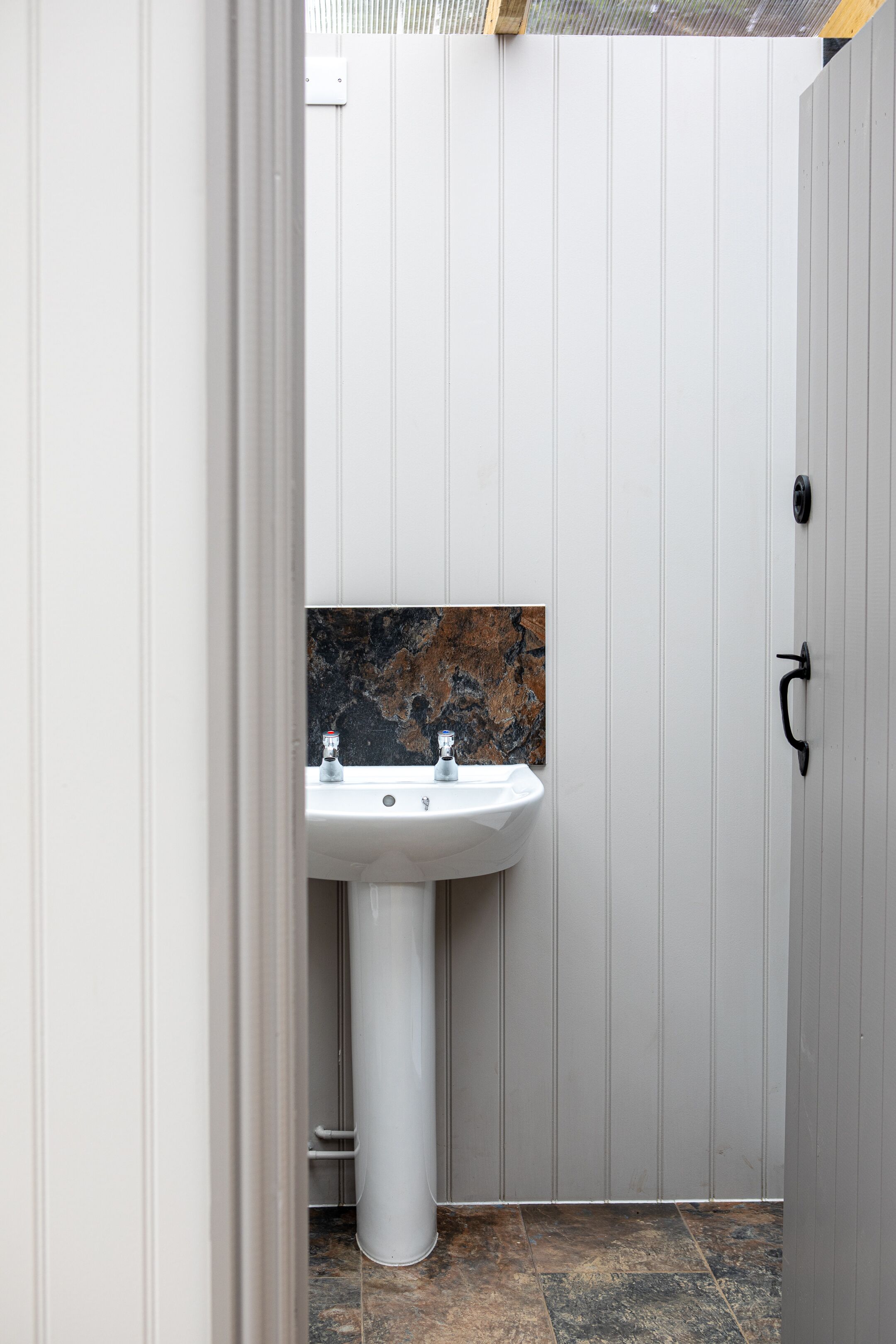 Een badkamer met een wit wastafeltje, een marmeren spatwand en een deur met een zwarte handgreep.