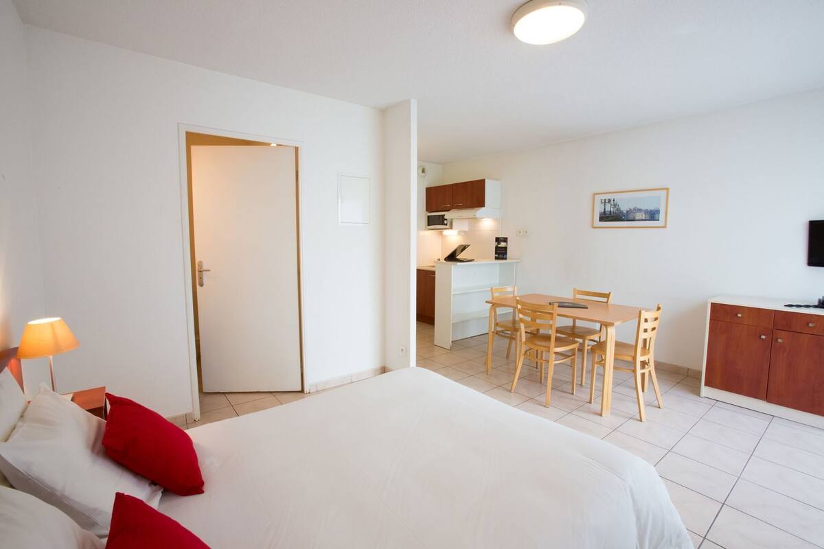 A hotel room with a bed, a desk, a chair, a dining table, a kitchen area, and a television.