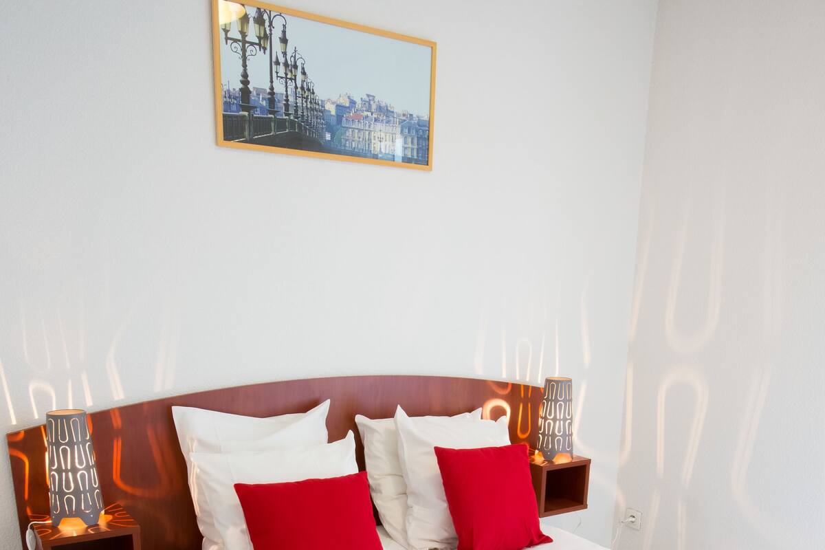 A neatly made bed with white and red pillows, a wooden headboard, and a cityscape painting above it.