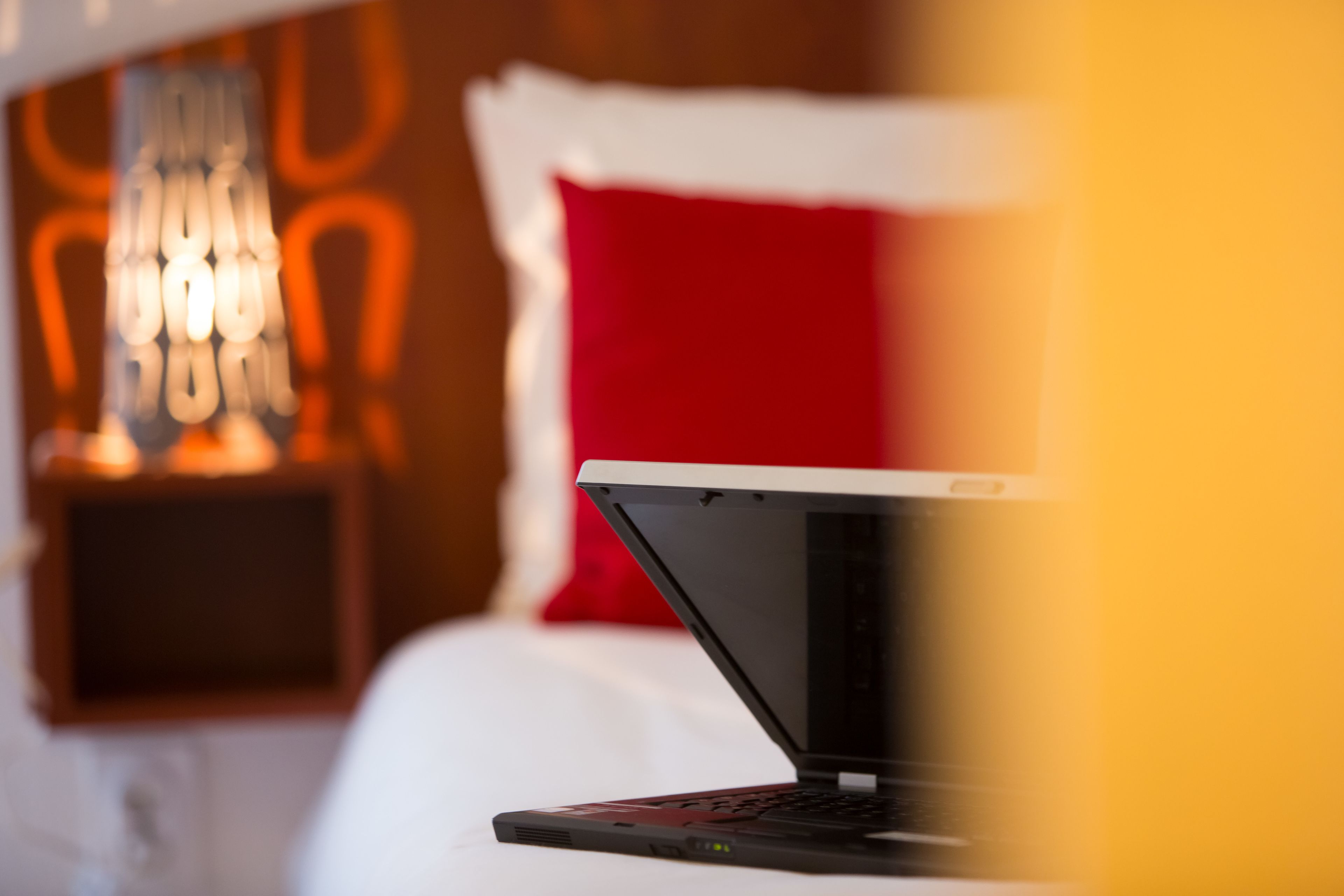 En bærbar computer på en seng i et hotelværelse med en rød pude og en vægmonteret lampe.