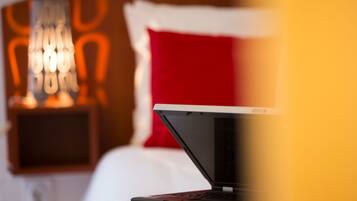 En bærbar computer på en seng i et hotelværelse med en rød pude og en vægmonteret lampe.