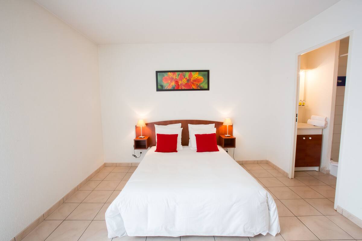 A hotel room with a large bed, two red pillows, a wooden headboard, two bedside lamps, a framed picture on the wall, and a bathroom visible through an open door.