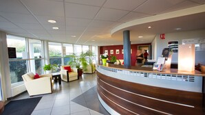 A modern reception area with a curved reception desk, comfortable seating, potted plants, and large windows providing natural light. - All Suites Appart Hôtel | Bordeaux - Mérignac Aéroport (Merignac)