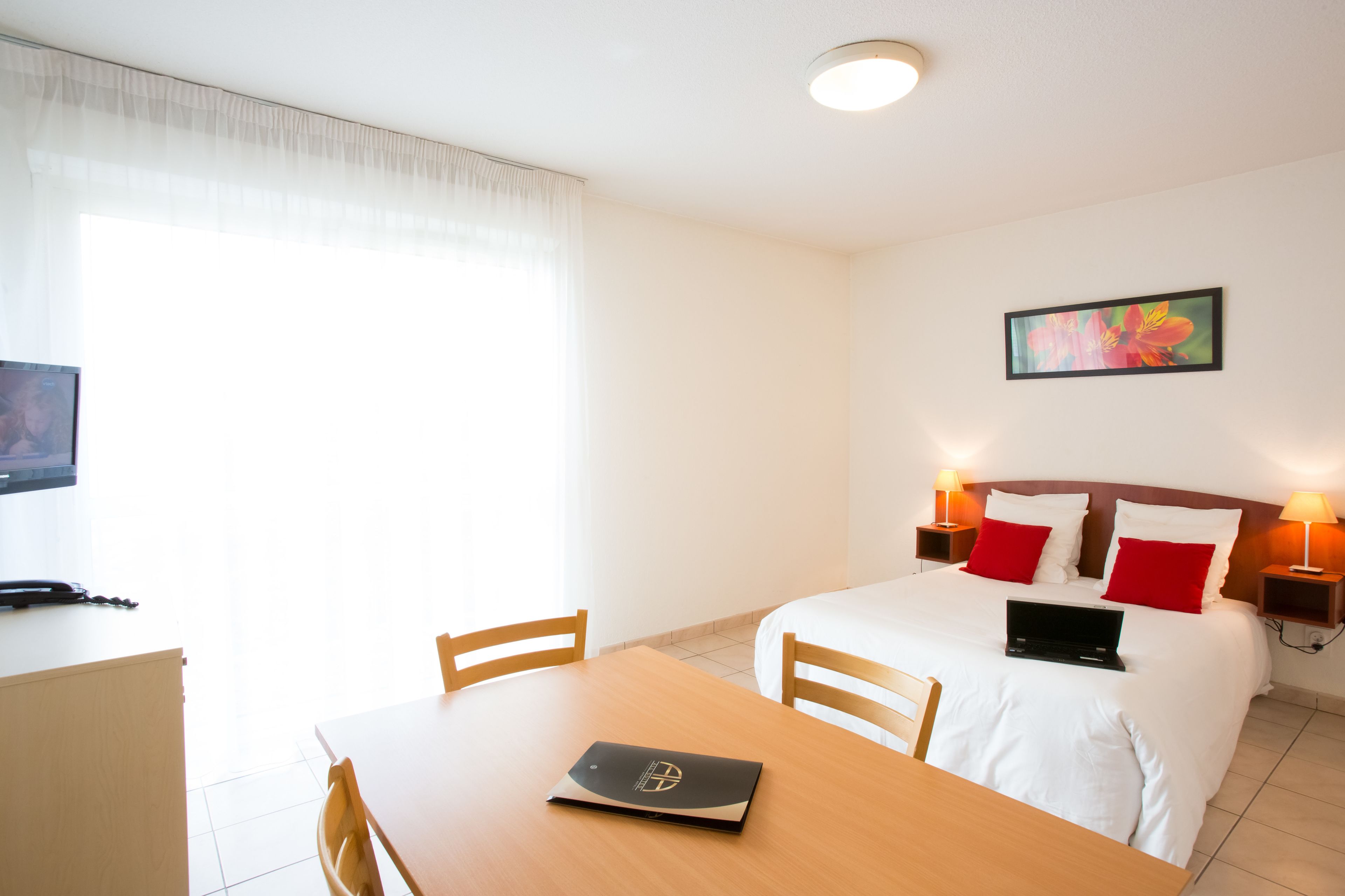 Habitación de hotel con cama, lámparas de noche, una laptop sobre la cama, un escritorio con una laptop y un televisor sobre un mueble.