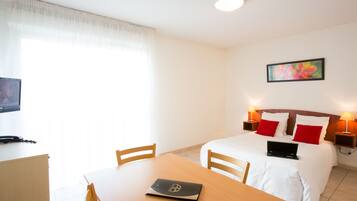 Habitación de hotel con cama, lámparas de noche, una laptop sobre la cama, un escritorio con una laptop y un televisor sobre un mueble.