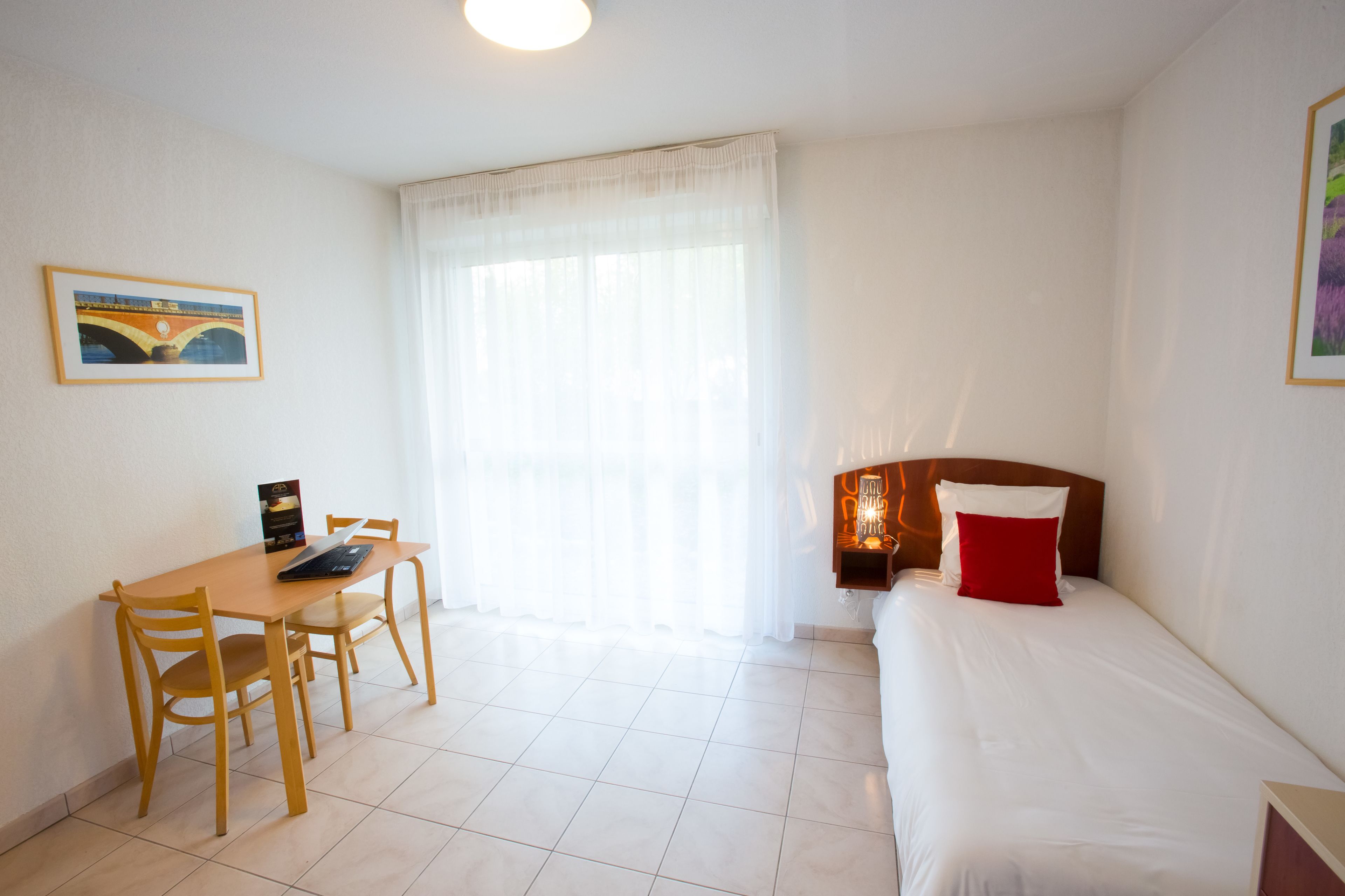 A small, well-lit room with a bed, a desk, and chairs. There is a window with sheer curtains, a framed picture on the wall, and a small table lamp on the bedside table.