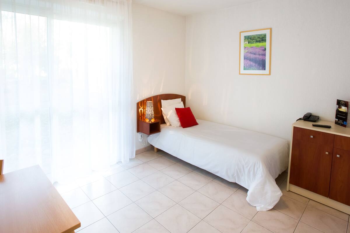 A hotel room with a bed, a nightstand, a TV, a desk, and a window with sheer curtains.