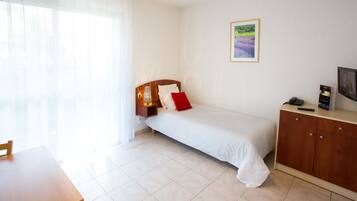 A hotel room with a bed, a nightstand, a TV, a desk, and a window with sheer curtains.