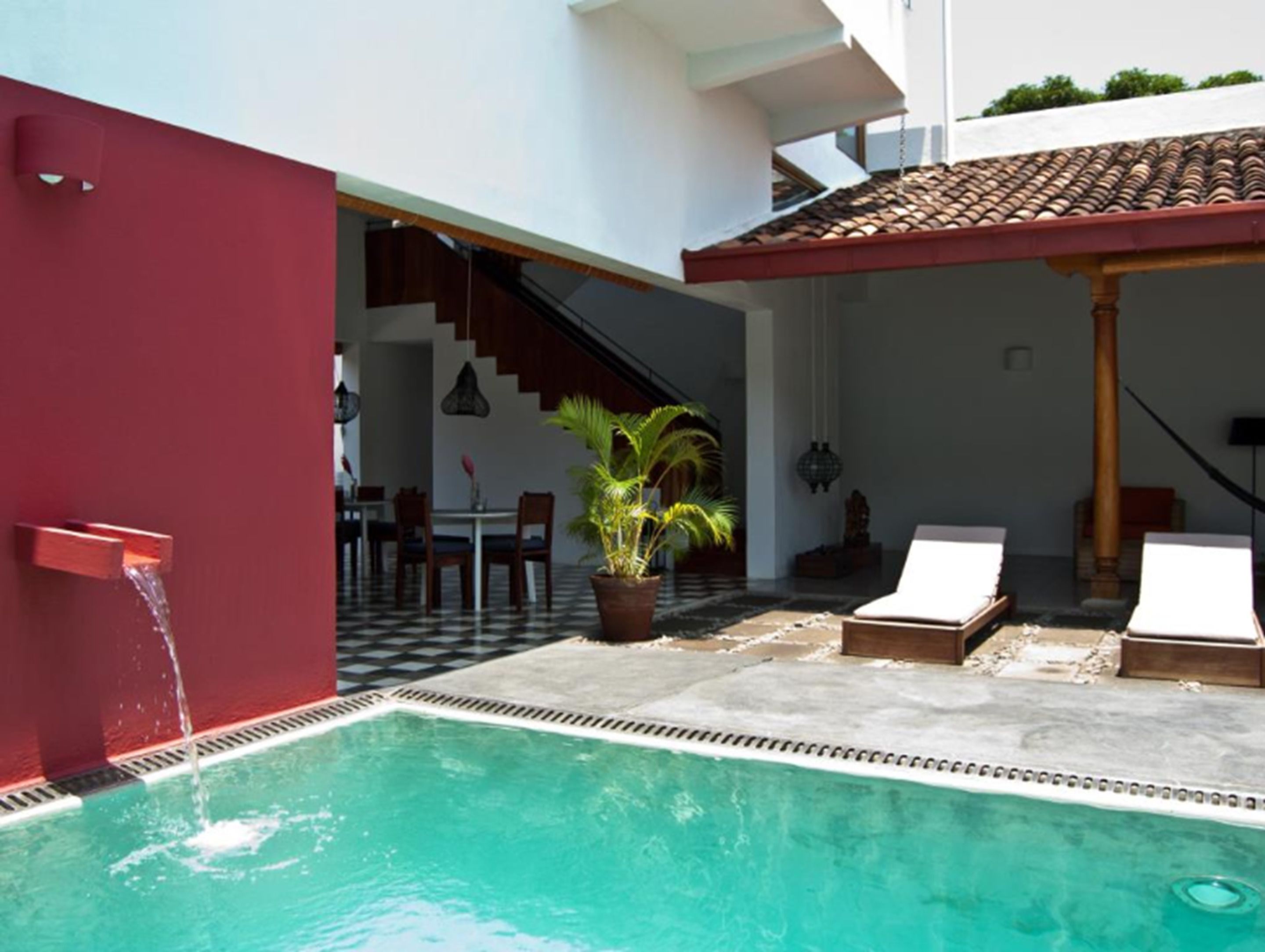 Una piscina con un muro rojo, una escalera que sube a un nivel superior y un comedor con sillas y una mesa.