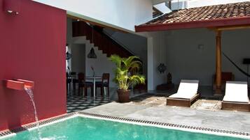 Una piscina con un muro rojo, una escalera que sube a un nivel superior y un comedor con sillas y una mesa.