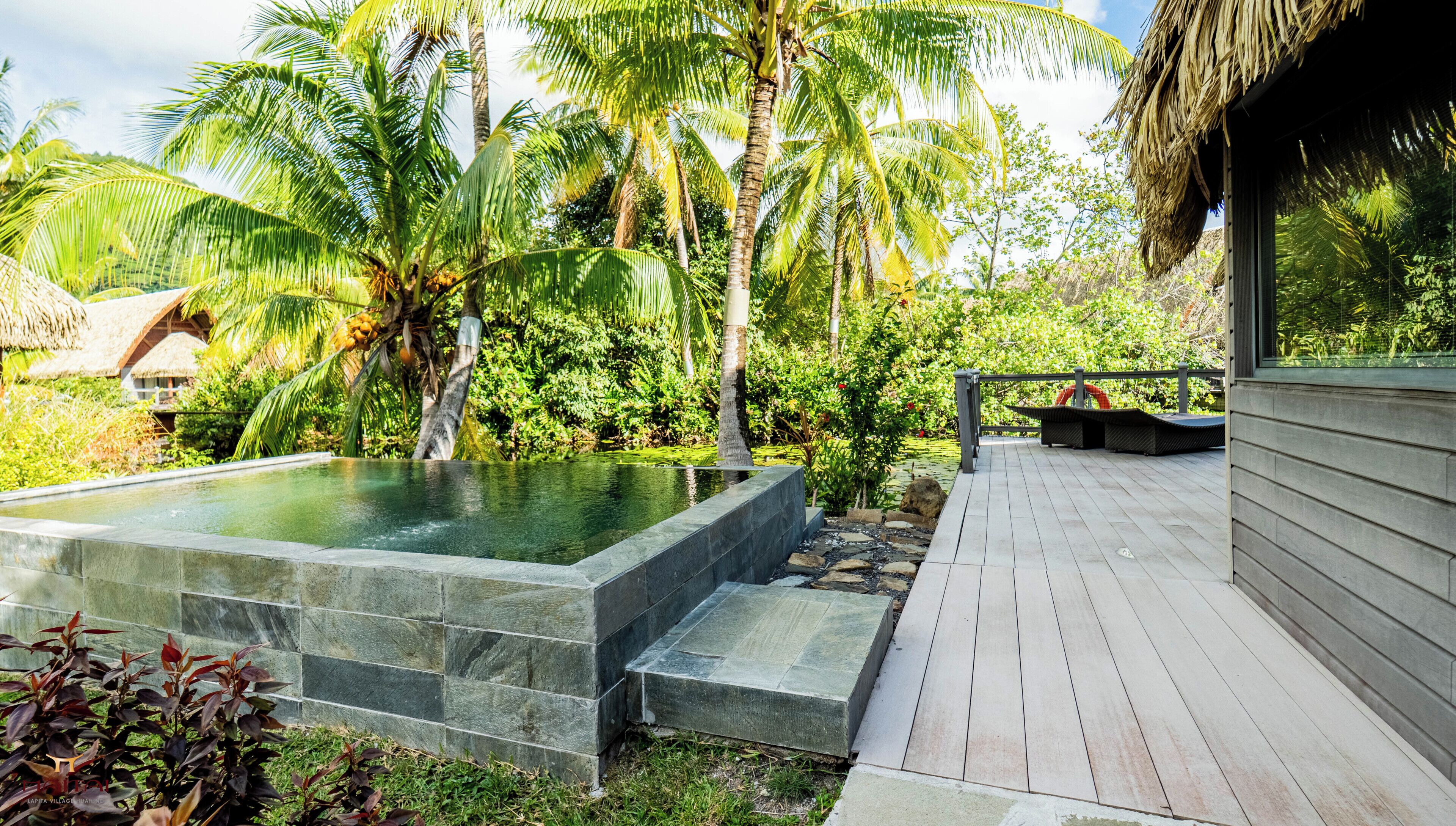 A hot tub surrounded by lush greenery, a wooden deck, and a thatched-roof structure.