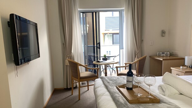Une chambre d’hôtel comprenant un lit, une télévision, une table à manger avec des chaises, un petit coin cuisine et un balcon avec des rideaux.