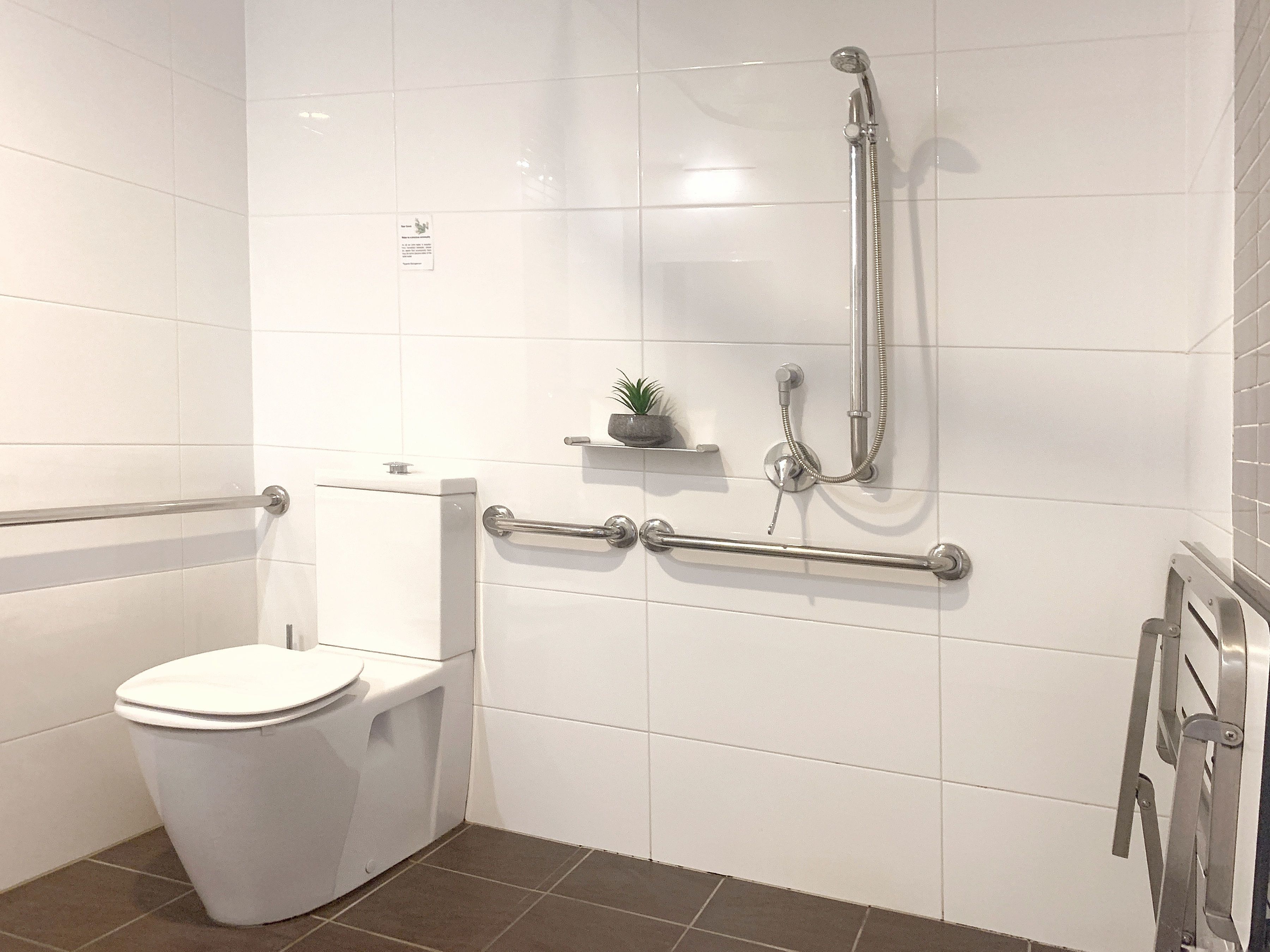 Un baño moderno con azulejos blancos, un inodoro, ducha con barras de apoyo y un estante pequeño con una planta.