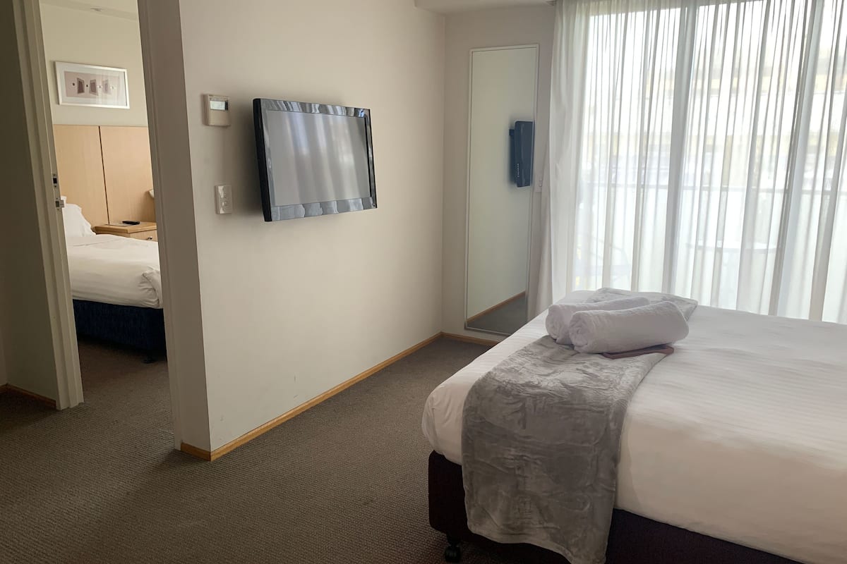 Quarto de hotel com cama, espelho, janela com cortinas e porta para outro aposento.