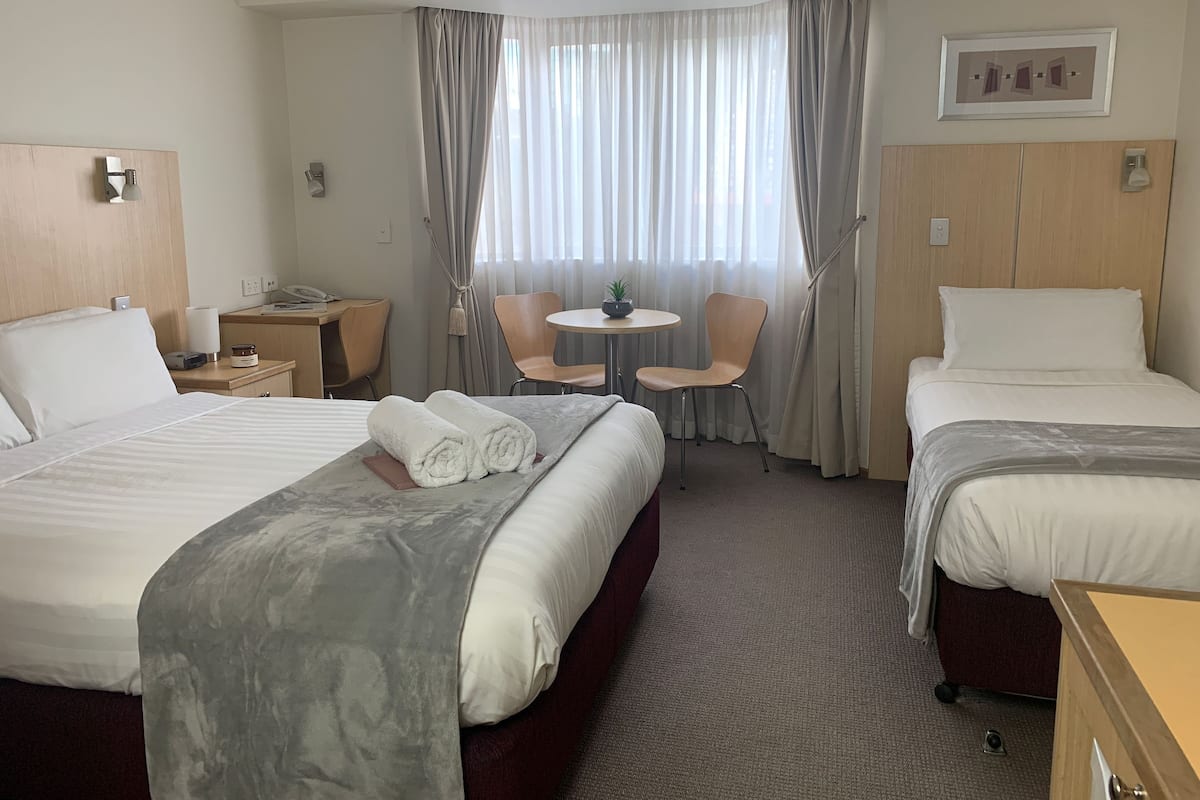 Quarto de hotel com duas camas, uma escrivaninha, uma cadeira, uma mesinha e uma janela com cortinas.