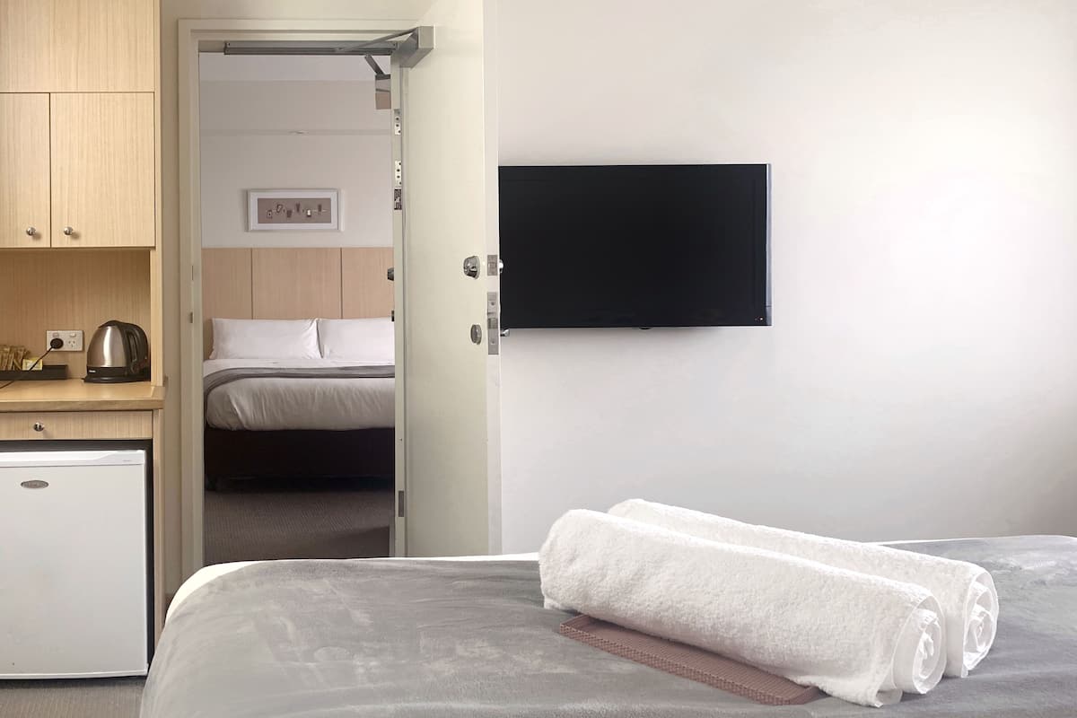 Quarto de hotel com cama, televisão na parede, pequeno espaço com cozinha equipado com chaleira e porta que dá para outro quarto.