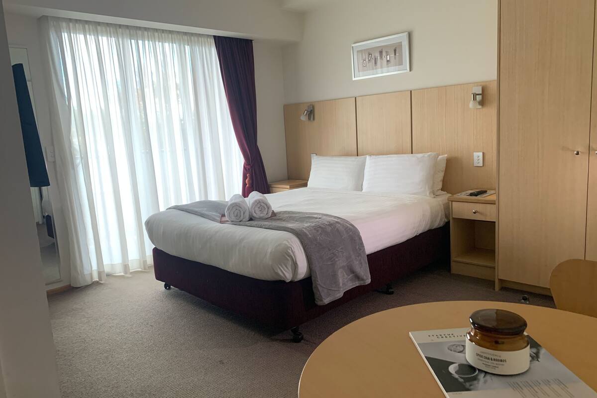 Quarto de hotel com cama, mesa de cabeceira, cadeira, mesa redonda com um livro e um pote, e janela com cortinas.